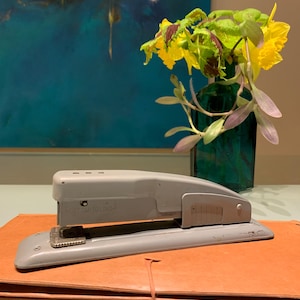 May include: A gray metal stapler with a black handle and a silver lever. The stapler is sitting on top of a brown leather folder. A vase with yellow flowers is in the background.