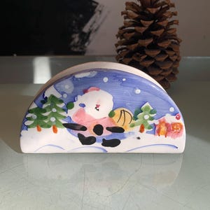 May include: A ceramic napkin holder shaped like a half-circle with a winter scene painted on it. The scene features a snowman in a pink hat and scarf, a blue sky with white snowflakes, and green pine trees.