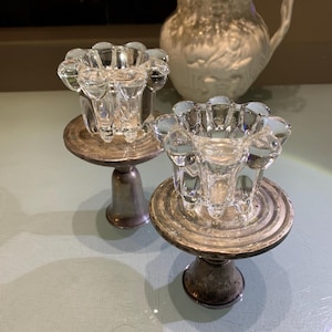 May include: Two clear glass candle holders with a flower-like design, set on silver-colored metal bases. A white pitcher with a floral relief pattern is in the background. The candle holders are a vintage style.