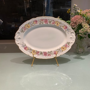 May include: A white oval serving platter with a gold rim and floral design. The platter is on a gold metal stand.