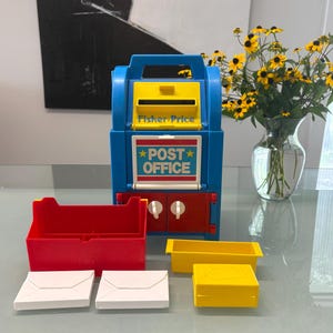 May include: A blue and yellow plastic Fisher-Price post office toy with a red base, featuring a mail slot, a sign reading POST OFFICE, and two white knobs. Included are a red bin, a yellow tray, a yellow box, and two white envelopes.