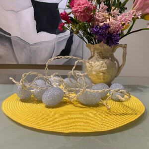 May include: A yellow woven placemat with a string of white fairy lights with silver glitter balls on top. The lights are arranged in a circle on the placemat. A white ceramic pitcher with a floral design holds a bouquet of pink, purple, and white flowers.
