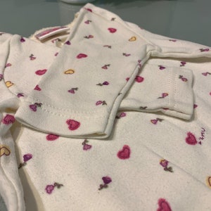 May include: Cream-colored baby outfit with a pattern of pink and yellow hearts and small purple flowers. The long-sleeved top has a wrap-around design. The cuffs of the sleeves are visible, and the fabric appears soft and comfortable.