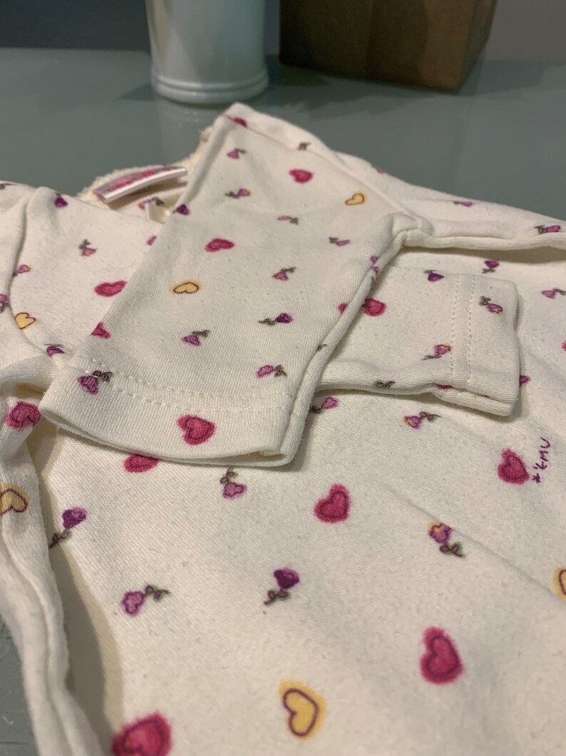 May include: Cream-colored baby outfit with a pattern of pink and yellow hearts and small purple flowers. The long-sleeved top has a wrap-around design. The cuffs of the sleeves are visible, and the fabric appears soft and comfortable.