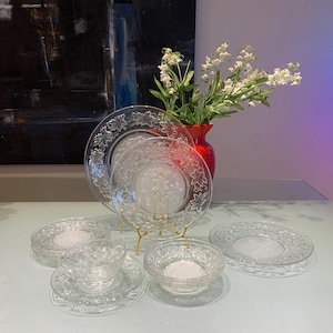 May include: A clear glass dinnerware set with a floral design. The set includes plates, saucers, bowls, and a teacup. A red vase with white flowers is in the background. The plates are displayed on a light blue surface.