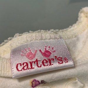 May include: Close-up of a white baby garment with a satin tag. The tag features two pink handprints above the brand name "Carter's" in pink lettering. A small white bow is visible.