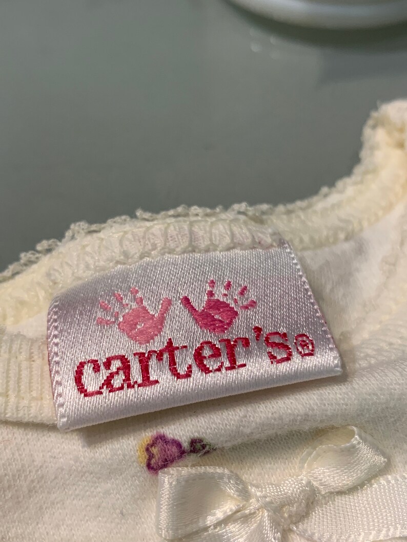 May include: Close-up of a white baby garment with a satin tag. The tag features two pink handprints above the brand name "Carter's" in pink lettering. A small white bow is visible.