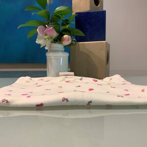 May include: A folded white baby onesie with a pattern of pink hearts and flowers. The onesie has a small bow at the neckline. A vase of flowers and decorative blocks are in the background.