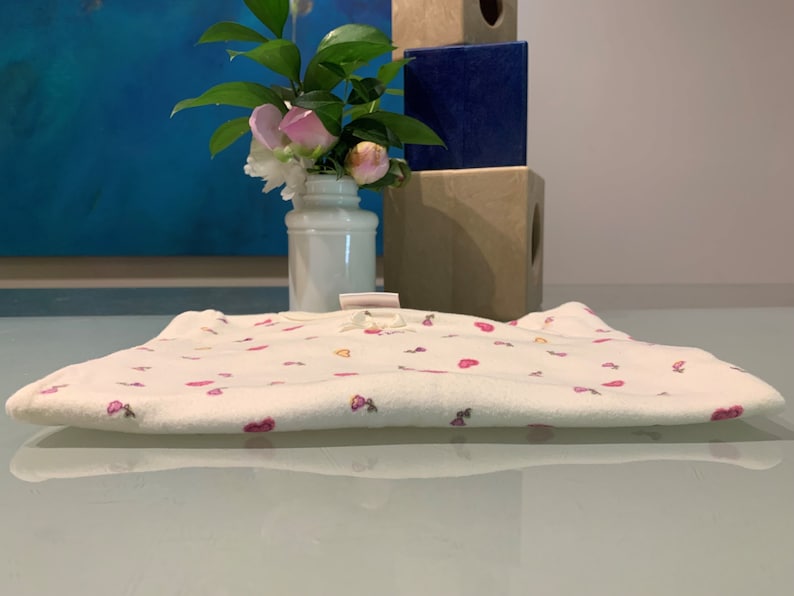 May include: A folded white baby onesie with a pattern of pink hearts and flowers. The onesie has a small bow at the neckline. A vase of flowers and decorative blocks are in the background.