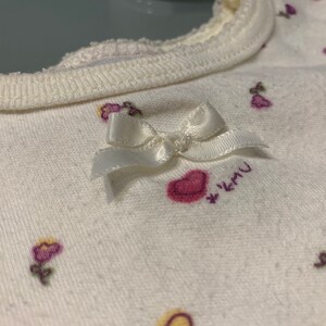 May include: Close-up of a white baby bib with a cream-colored trim. The bib is decorated with small pink hearts and purple flower designs. A small white bow is attached to the bib.