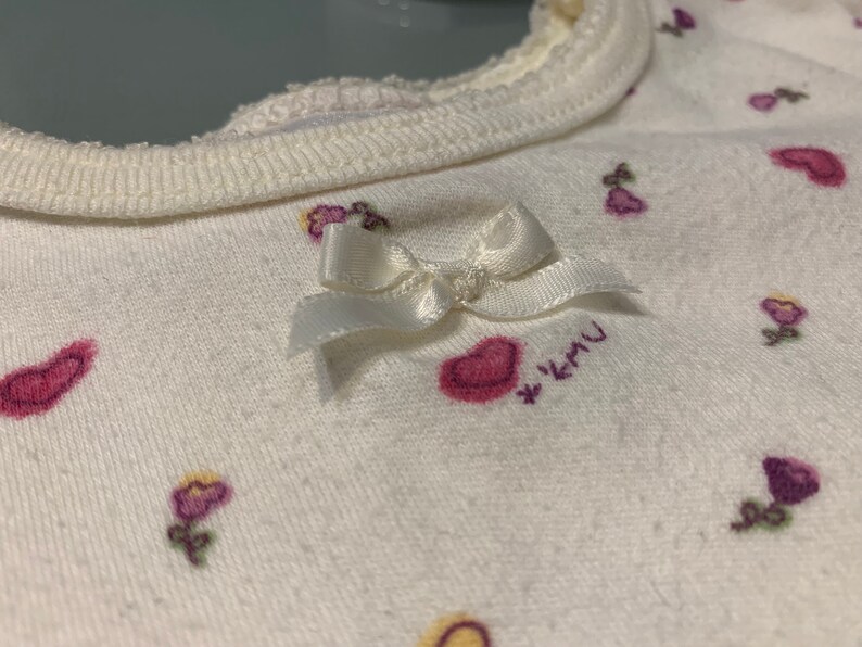 May include: Close-up of a white baby bib with a cream-colored trim. The bib is decorated with small pink hearts and purple flower designs. A small white bow is attached to the bib.