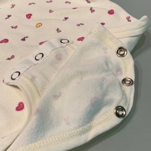 May include: Close-up of a white baby onesie with a pattern of pink and purple hearts and floral designs. The onesie has a snap closure and a soft, textured fabric. The edges are finished with a white trim.