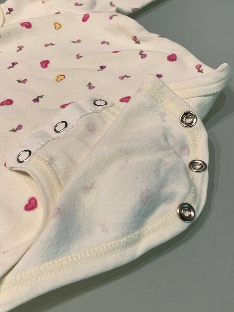 May include: Close-up of a white baby onesie with a pattern of pink and purple hearts and floral designs. The onesie has a snap closure and a soft, textured fabric. The edges are finished with a white trim.