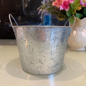 May include: A galvanized metal bucket with two arched handles. The bucket has a speckled, textured surface and a cylindrical shape. The background includes a vase of pink flowers and a dark abstract painting.