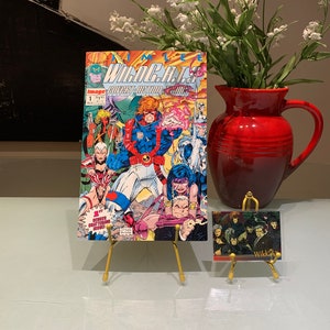 May include: A comic book titled "WildC.A.T.s" by Jim Lee, issue #1, August 1992, with a cover featuring a group of superheroes in action poses. The comic book is displayed on a gold metal stand. A WildC.A.T.s trading card is displayed on a similar stand.