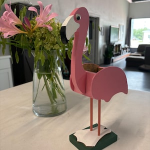 May include: A pink wooden flamingo figurine with a white and black beak, a googly eye, and two thin coral-colored legs standing on a rectangular green base with white webbed feet. The flamingo has a hollow opening on its back.