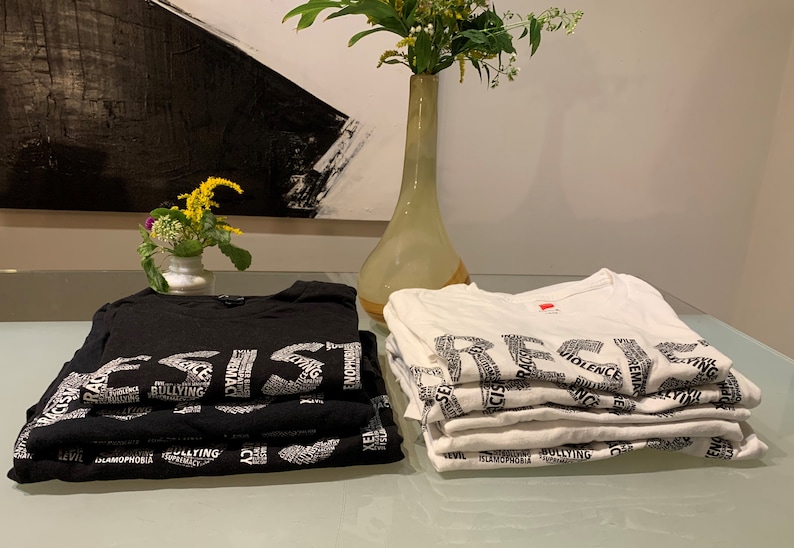 May include: A stack of black t-shirts with the word "RESIST" spelled out in white letters, and a stack of white t-shirts with the word "RESIST" spelled out in black letters. The shirts are made of a soft, comfortable material and feature a variety of words related to social justice and resistance.