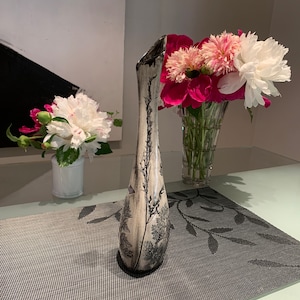 May include: A white ceramic vase with a black floral design. The vase is tall and slender with a narrow opening. The vase is sitting on a black and white placemat.