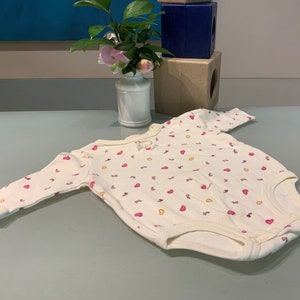 May include: A cream-colored baby bodysuit with long sleeves, adorned with a pattern of small pink and yellow hearts. The bodysuit has a round neckline and a small bow detail. The garment is made of soft fabric.
