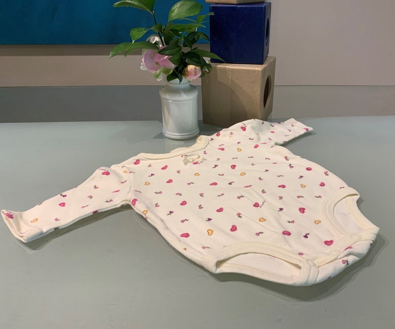 May include: A cream-colored baby bodysuit with long sleeves, adorned with a pattern of small pink and yellow hearts. The bodysuit has a round neckline and a small bow detail. The garment is made of soft fabric.