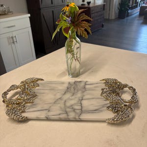 May include: A rectangular white marble serving tray with grey veining, featuring ornate metallic handles shaped like clusters of grapes and vine leaves on both ends. The tray is positioned horizontally on a flat surface with a clear glass vase.