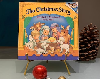 May include: A vintage children's book titled "The Christmas Story" featuring Ruth J. Morehead's Holly Babes. The cover depicts a colourful illustration of the nativity scene with animals and a manger.