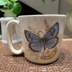 May include: White ceramic mug with a blue butterfly design. The butterfly has black markings and is surrounded by green leaves and flowers.