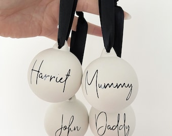 Peut inclure: Quatre boules de Noël en céramique blanche avec des rubans noirs. Les boules sont personnalisées avec les noms "Harriet", "Mummy", "John" et "Daddy" en écriture noire.