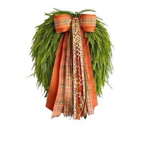 May include: A teardrop-shaped wreath made of vibrant green foliage, featuring a large decorative bow at the top. The bow is crafted from orange and green plaid ribbon, with long, trailing ribbons in solid orange, plaid, and leaf-patterned fabric.