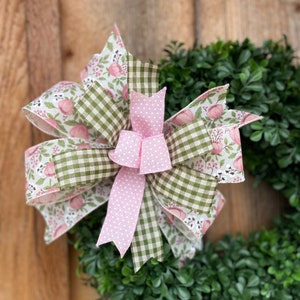 May include: A decorative wreath featuring a large bow. The bow is made of ribbons with floral, gingham, and polka dot patterns in pink, green, and white. The wreath base is composed of green foliage, set against a wooden background.