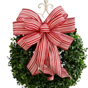 May include: A red and white striped bow is tied around a green boxwood wreath. The bow is made of a satin ribbon and has a large, full shape.