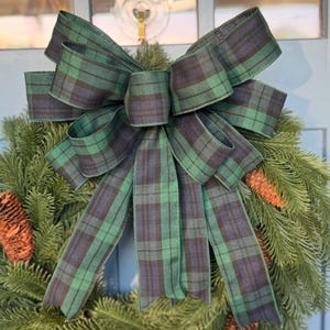 May include: A Christmas wreath with a large plaid bow in shades of green and navy blue. The wreath is made of evergreen branches and decorated with pine cones. The bow is the focal point, adding a festive touch to the holiday decor.