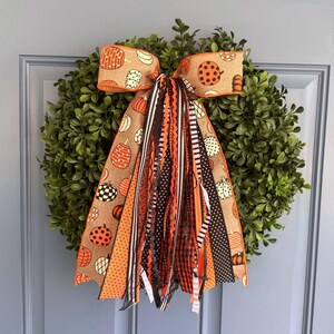 May include: A decorative wreath featuring a large bow made of tan burlap ribbon with a pumpkin print. The bow has long, dangling streamers in various patterns including black and white stripes, orange polka dots, and solid black and orange ribbons.