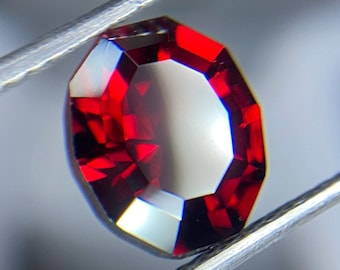 May include: A deep red gemstone with a faceted octagonal shape, held by tweezers.