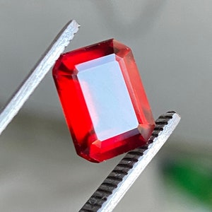 May include: A red gemstone with a rectangular shape is held by a pair of tweezers.