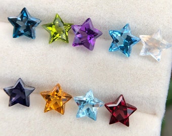 May include: A display of eight star-shaped gemstones in a white velvet box. The gemstones are arranged in a row and are of various colors, including blue, green, purple, orange, and red.
