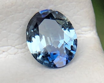 Könnte beinhalten: Ein oval geschliffener Edelstein mit einem Farbverlauf von Blau, von hell bis tief Indigo. Der Stein liegt auf einer weißen Oberfläche, mit einem verschwommenen grünen Hintergrund. Die Facetten des Steins fangen das Licht ein und erzeugen einen funkelnden Effekt.