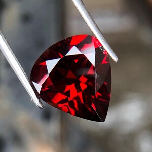 May include: A deep red gemstone with a triangular cut, held by tweezers. The gemstone is faceted and reflects light.