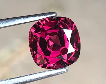May include: A faceted, cushion-cut gemstone in a vibrant shade of red, held by silver tweezers. The gem's multiple facets reflect light, creating a sparkling effect. The background is a soft, blurred neutral tone.