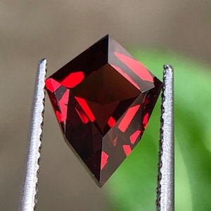 May include: A square-cut, deep red gemstone held by silver tweezers. The gem's facets reflect light, displaying its rich colour and clarity. The background is blurred, highlighting the gemstone.