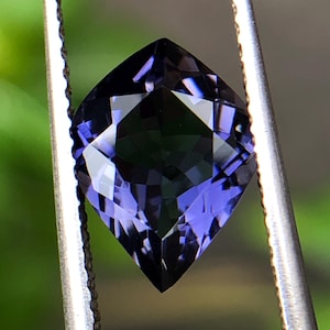 以下が含まれることがあります： 金属製のピンセットで保持された、クッションカットのファセット宝石。 宝石は深い紫がかった青色で、光を反射する複数のファセットがあります。 背景は柔らかくぼやけた緑色で、自然な葉を連想させます。