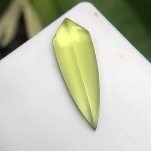May include: A translucent, pale lime-green gemstone cut into a sharp, elongated shield or leaf-like shape. The stone features a distinct central ridge running vertically, creating two symmetrical, faceted slopes that taper to a pointed tip.