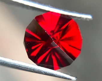 Puede incluir: Una piedra preciosa de color rojo intenso con un corte facetado, sostenida por unas pinzas. La piedra preciosa tiene un patrón de estrella.