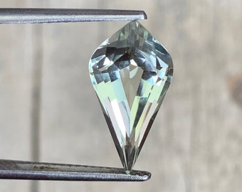 May include: A light green gemstone with a pointed, teardrop shape. The gemstone is held in place by a pair of tweezers.