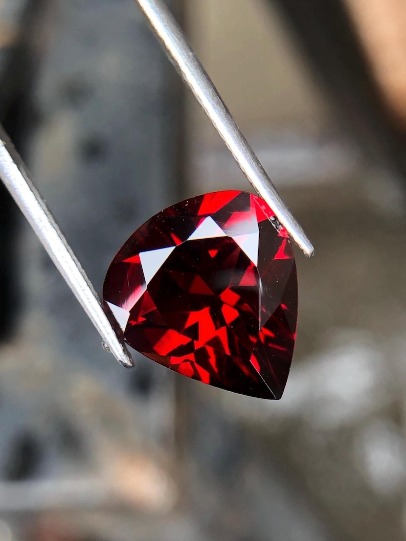 May include: A deep red gemstone with a triangular cut, held by tweezers. The gemstone is faceted and reflects light.