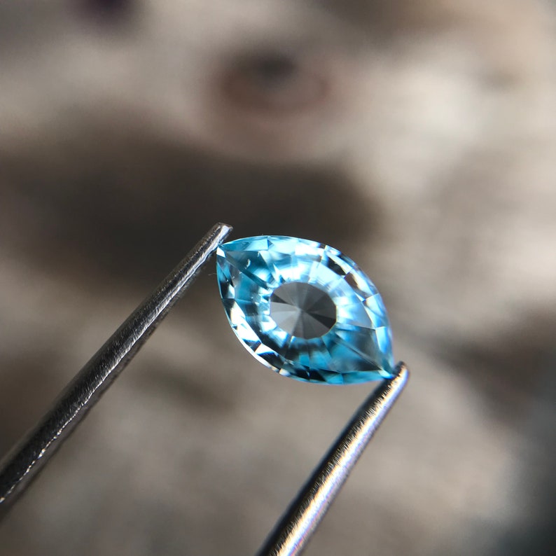 May include: A light blue marquise-shaped gemstone with a faceted design. The gemstone is held in tweezers.