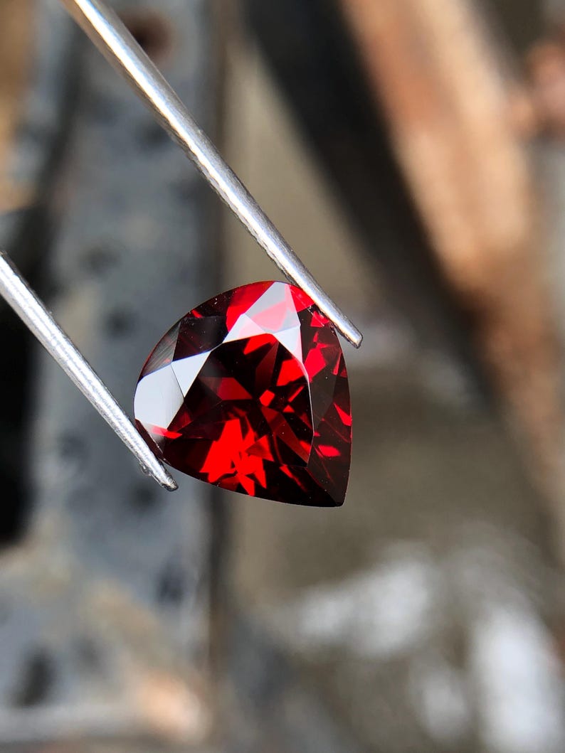 May include: A deep red heart-shaped gemstone with facets, held by tweezers.