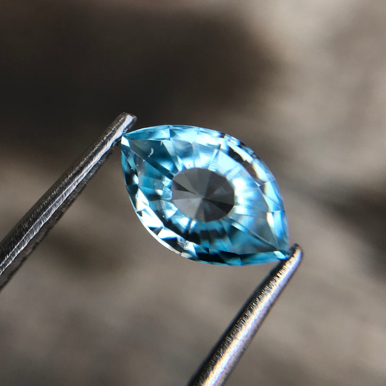 May include: A light blue marquise-shaped gemstone with a faceted design. The gemstone is held in a pair of tweezers.