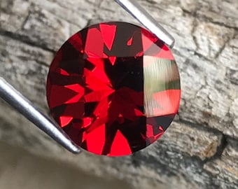 May include: A round, faceted, deep red gemstone held by silver tweezers. The gem's surface reflects light, highlighting its internal structure. The gemstone is set against a textured, grey wooden surface.