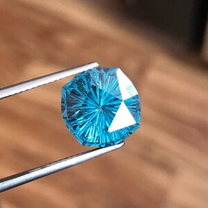 Puede incluir: Una piedra preciosa azul con un patr&oacute;n de estrella, sostenida en unas pinzas. La piedra preciosa est&aacute; facetada y tiene una forma hexagonal.